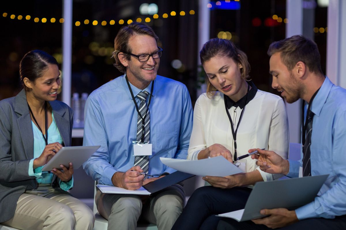 Cuatro profesionales con acreditaciones sentados revisando documentos y tablet en reunión de convención en exterior nocturno con luces de fondo